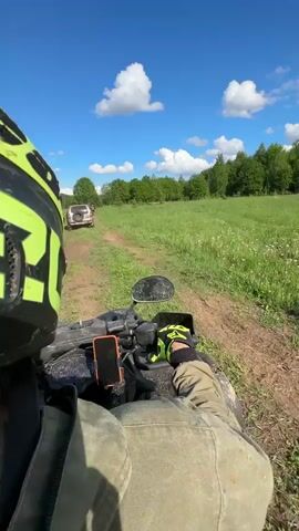 The video shows the first-person perspective of someone riding an ATV on a dirt path through a grassy field. The rider is wearing a green and black helmet and Fox gloves. In front of the ATV, another vehicle, possibly a Jeep, is traveling along the same path. The sky is blue with scattered white clouds, and there's a treeline in the background. The grass is green and lush, and the path appears muddy in places. The sun is shining, creating a bright, outdoor scene. The camera moves smoothly as the ATV travels, providing a clear view of the environment.