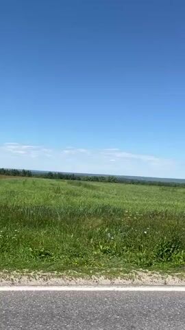 The video shows a view from inside a moving vehicle. The camera is pointed outwards, capturing a rural landscape under a clear blue sky with scattered white clouds. The landscape consists of rolling green fields, dotted with some trees and utility poles. The foreground shows a portion of the road and the side mirror of the vehicle. The side mirror reflects the road ahead and the passing scenery, including a speed limit sign with a graphic of a car. The video appears to be shot during the daytime.
