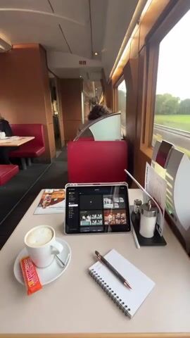 The video shows a close-up, eye-level, static shot of a table in what appears to be a train dining car. On the table, a white cup of coffee with latte art sits on a saucer, accompanied by an orange-wrapped candy bar. A tablet, displaying a grid of images, is positioned in the center of the table, angled slightly towards the viewer. To the right of the tablet, a small notepad with a pen rests open, and a salt and pepper shaker set is visible. In the background, through a large window, a blurred landscape of greenery and tracks suggests the train is in motion. The interior of the train car is visible, with red cushioned seating and wooden paneling. A person's head and shoulders are partially visible in the background, seated in the red seating. The overall lighting is soft and natural, coming from the window and interior lighting.