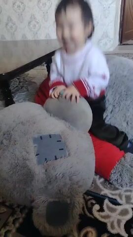 A young child wearing a white sweater with red trim and dark pants is sitting on a large, gray stuffed teddy bear with a gray patch on its back. The child is smiling and appears to be laughing. The teddy bear is positioned on a gray, furry seat cushion with red pillows underneath. The scene takes place on a patterned rug in front of a wall with decorative wallpaper. A dark brown table with a black, shiny top is visible in the background. The child's movements are slightly blurred, indicating motion.