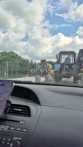 The video, filmed from inside a car, shows a construction site. Several people, dressed in work clothes, are gathered around a yellow cement mixer truck. One person appears to be operating the mixer, while others are working nearby. A blue tractor is parked behind the cement mixer. The scene is outdoors with trees and a cloudy sky visible.