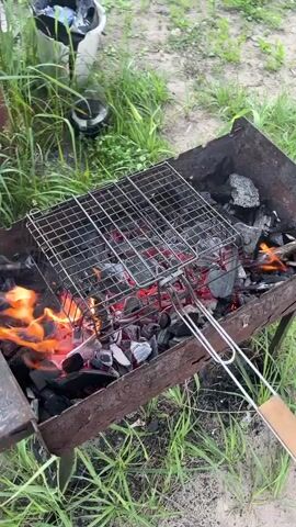 A close-up shot shows a barbecue grill filled with glowing red coals and flames. A metal grill rack is positioned above the coals. A person wearing a green glove enters the frame from the right, holding a light blue cutting board, and uses it to fan the coals, creating a cloud of smoke. The background consists of green grass and dry ground, suggesting an outdoor setting.