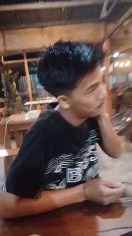 A young man with dark, spiky hair and a light mustache is seated at a wooden table in what appears to be a semi-outdoor restaurant or cafe. He is wearing a black t-shirt with a white design that includes the letters "BBS" (or "BSS"). The man is actively wiping his face, specifically his cheeks and around his eyes, with a white tissue or wipe. He holds another piece of the tissue in his left hand. The setting features wooden pillars, a corrugated roof, and other tables and chairs in the background, suggesting an evening or nighttime scene due to the dim lighting and visible artificial lights. The overall atmosphere seems casual.