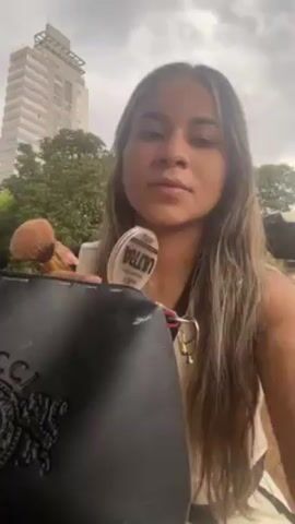 A young woman applies makeup using a brush and compact in an outdoor setting. She holds a black Gucci purse with her left hand while using her right hand to apply makeup to her face. She is wearing a white top with black trim. Trees and a tall building are visible in the background. The sky is overcast.
