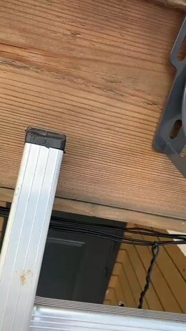 A close-up shot shows a person using a cordless drill to attach a gray plastic bracket to a wooden surface. The drill bit is actively screwing into a pre-drilled hole, securing the bracket. The bracket appears to be part of a larger system, possibly for holding pipes or wires. The wooden surface is made of horizontal planks with visible grain and texture. A silver ladder is partially visible in the lower left corner of the frame, with black tape covering the end of one of its rungs. Black wires are strung horizontally beneath the wooden surface. The overall lighting suggests an outdoor environment.