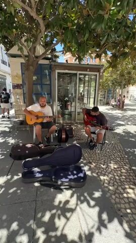 Two men are playing acoustic guitars on a sunny day, seated on chairs in front of a small kiosk. They are positioned on a cobblestone sidewalk lined with trees that cast dappled shadows. Three guitar cases are stacked on the ground in the foreground. The man on the left is wearing a white t-shirt and shorts, while the man on the right is wearing a dark t-shirt and shorts. In the background, other people are walking on the sidewalk, and buildings are visible. The overall setting appears to be a public outdoor space, possibly a street or plaza.