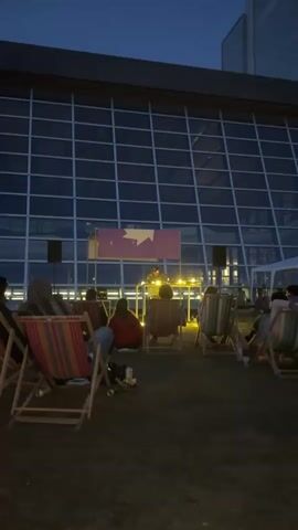 The video shows an outdoor event at dusk. A DJ is set up on a platform, with two speakers on either side. A large banner with a purple background and a yellow starburst design is behind the DJ. A crowd of people are seated in striped deck chairs, facing the DJ. The background consists of a large glass building with a dark sky overhead. The atmosphere appears to be relaxed and casual, with people enjoying the music.