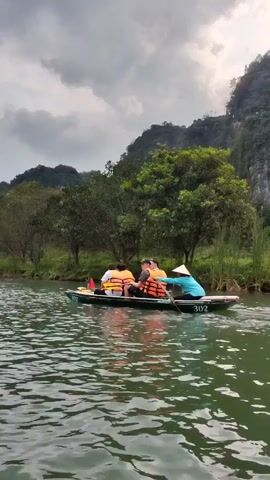 The video depicts two boats traversing a river. Each boat is occupied by at least three individuals, all of whom are wearing life vests. One of the occupants in each boat is wearing a conical Asian hat and using oars to propel the boat forward. The river is surrounded by lush green trees and mountains, under a cloudy, overcast sky.