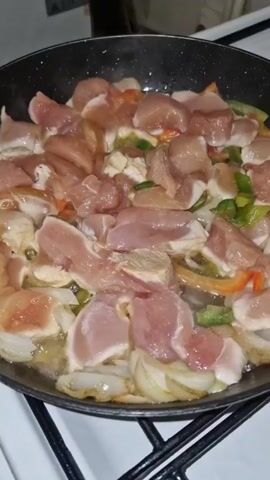 A close-up shot shows a black frying pan on a white stovetop. The pan contains raw, cubed chicken pieces along with sliced onions, green bell peppers, and a few pieces of orange bell pepper. The ingredients are being cooked in oil or butter, visible as a shimmering liquid in the pan. The stovetop has black grates. The scene is domestic, likely a kitchen during meal preparation. The camera angle is slightly angled towards the ingredients in the pan. Audio suggests people are talking in the background.