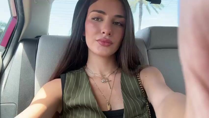 A young woman with long dark hair is sitting in the backseat of a car. She is wearing a green pinstripe vest over a black top and several necklaces. She is looking at the camera and appears to be taking a selfie. The car's interior is visible behind her, including the headrest of the seat in front and part of the back seat. Sunlight is coming through the windows, and palm trees can be seen outside. The woman's right hand is partially visible in the foreground, suggesting she is holding the camera.