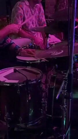 A drummer with pink drumsticks plays a drum set in a dimly lit room. The drummer is wearing a colorful patterned shirt. The drum set includes a snare drum and cymbals. The room is lit with pink and green lights.