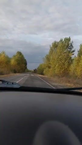 The video, captured from inside a vehicle, shows a view through the windshield as the car travels down a two-lane asphalt road. The road is in fair condition, with some visible cracks and wear. Trees with autumn foliage in shades of yellow and green line both sides of the road. The sky is overcast with scattered clouds. The foreground shows the dashboard of the car, including the base of the windshield wipers. There are no people or other vehicles visible in the scene. The lighting suggests it is daytime, likely late afternoon or early morning due to the soft light.