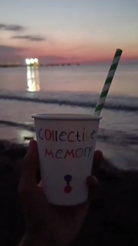 The video shows a close-up of a hand holding a white paper cup with a green and white striped straw. The cup has the words "COLLECTIVE MEMORY" written on it in colorful letters, and a small blue and red logo at the bottom. The background is a sunset over the ocean, with a pier or walkway visible in the distance. The sky is a gradient of pink, orange, and purple. The water is calm with gentle waves. The overall mood is peaceful and serene.