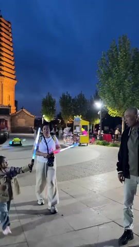 The video shows a family in a public square with a tall, lit-up pagoda-like structure in the background. A woman in a white shirt and white pants is holding two light sabers. A young child in a tan coat is holding one of the light sabers, and an older man in a black jacket and grey pants is standing nearby. The square is paved with square tiles and has trees and streetlights. People are walking around in the background. There is a vendor stand to the right, and several illuminated decorative pieces on the ground. The sky is dark blue, indicating it is dusk or nighttime. The focus of the video appears to be the family interacting with the light sabers in the square.