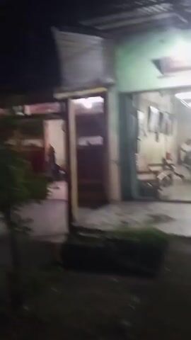 A night-time, shaky, first-person perspective video shows an exterior view of buildings. The camera pans across a row of attached structures, some with red doors and what appear to be windows. A car is partially visible in the foreground, and some green foliage is present. Lights illuminate the buildings, casting shadows. The scene appears to be a residential or commercial area.