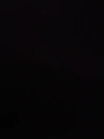 The video opens to complete darkness, slowly revealing a very dimly lit outdoor scene at night. A few faint, distant light sources are visible, appearing as blurry white points against the dark background, possibly from streetlights or distant buildings. In the foreground, a small group of 2-3 people can be discerned, illuminated by a very low, localized light source, perhaps a flashlight or a phone screen, casting a small glow on the ground around them. Their figures are mostly silhouettes, and specific details or activities are unclear due to the extreme low light conditions. The setting appears to be an open area or pathway, but no distinct features or landmarks are identifiable. The sky is dark, suggesting clear weather. No text is visible.