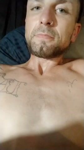 Here is a concise description of the video: 

The video shows a shirtless man lying down. He has tattoos on his chest and abdomen, a short beard, and a nose ring. He is looking towards the camera.  The background is indistinct, showing bedding in a dark color.