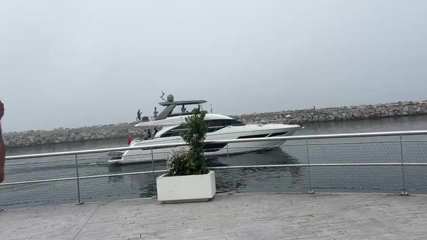 A white motor yacht is moving through a channel of water. The yacht has several people on its deck. The water is calm and reflects the overcast sky. On either side of the channel are stone jetties. In the foreground, there is a paved walkway with a metal railing and a planter containing a small tree and flowers. The overall scene suggests a hazy or foggy day.