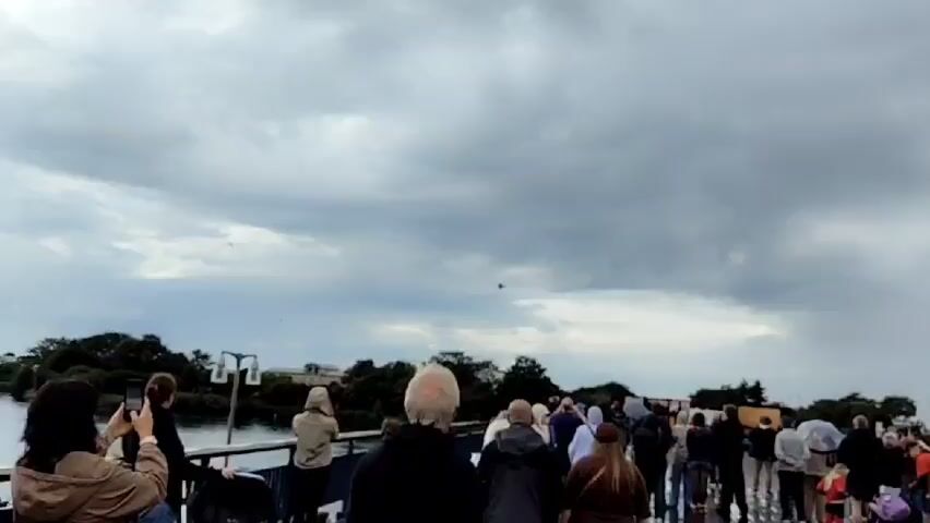 The video shows a crowd of people standing on what appears to be a bridge or elevated walkway, looking up at the sky. The sky is overcast with dramatic, dark clouds, and there is a hint of rain or a storm in the distance. Several birds are visible in the sky, and at least one aircraft, possibly a fighter jet, is captured flying at a distance. The people are dressed in casual clothing, and some are holding umbrellas. The overall atmosphere suggests a moment of shared observation of an aerial event, possibly an air show or a passing aircraft, taking place under threatening weather conditions.