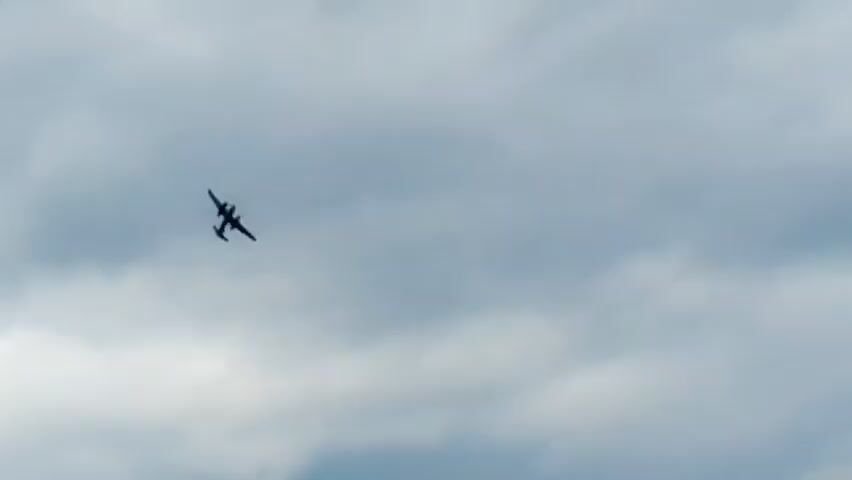 A small, dark twin-engine airplane flies against a cloudy sky. The airplane is silhouetted against the overcast background, with its wings and tail clearly visible. It appears to be moving across the frame from left to right, slightly angled downwards. The clouds are grey and diffuse, indicating an overcast or partly cloudy day. There are no other objects or people visible in the frame. The focus is entirely on the airplane and the sky.