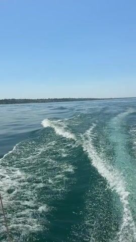 This is a first-person perspective video shot from a boat traveling on a body of water. The scene is dominated by the deep blue water, with white foam and waves trailing behind the boat, indicating movement. The sky above is a clear, bright blue, suggesting a sunny day. In the distance, a long, low-lying shoreline is visible, covered with greenery and some structures, possibly indicating land or an island. The overall impression is of a peaceful and scenic boat ride on a clear day, possibly in a coastal area or a large lake. The time of day appears to be daytime, likely late morning or early afternoon due to the bright sunlight and clear sky. No people are directly visible in the frame. The video captures the motion of the boat and the dynamic interaction of the water, creating a sense of journey and exploration. The location cue of "Lazise, Italy" places this scene on Lake Garda, known for its picturesque towns and clear blue waters, which aligns with the visual elements presented in the video. The mood conveyed is serene and enjoyable, consistent with a recreational boat trip on a pleasant day. There is no visible text in the video.