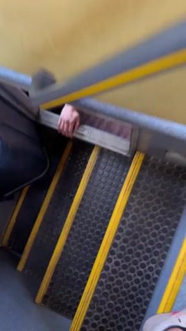 A person is sitting in a compartment. Their hand is visible on the side of the compartment, and their legs are inside. The person is wearing white sneakers with green laces, white socks, and shorts. The compartment has a brown carpet and a gray frame. The view is looking up from a staircase with yellow strips and black treads.