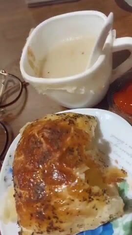 The video shows a close-up view of a meal. In the foreground, a golden-brown pastry with visible herbs is on a white plate. A bite has been taken out of the pastry, revealing a filling. To the right of the pastry is a small container of red sauce, possibly ketchup. In the background, a white mug with a handle contains a light-colored soup with a spoon. The mug appears to have a textured rim. A pair of eyeglasses with brown frames is visible on the left side of the image. The setting appears to be a table with a wooden surface. The lighting is dim, suggesting an indoor environment. The video focuses on the food items, highlighting their textures and colors.