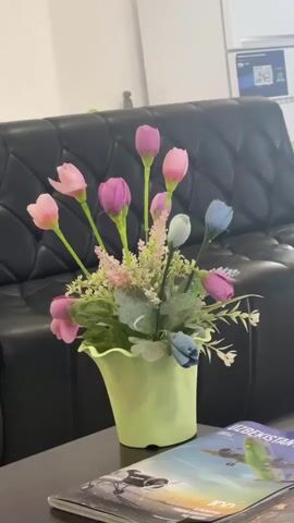 The video shows a close-up shot of a bouquet of artificial flowers in a light green vase, positioned on a dark brown coffee table. The flowers are predominantly pink and purple tulips, with a few blue and white accents. The bouquet is set against a blurred background of a black leather couch and a white wall. To the right of the vase, a stack of magazines is visible on the coffee table, with the top magazine featuring an image of an airplane and the word "Uzbekistan." The camera is stationary, and the focus shifts slightly throughout the clip, blurring the background and some of the foreground elements. There are no people visible in the video, and no actions or interactions are depicted. The lighting is bright, suggesting an indoor environment.