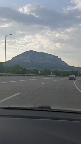 The video shows a view from inside a car driving on a highway. The highway has multiple lanes and is bordered by a guardrail. Beyond the guardrail, there are trees and a large mountain in the background. The sky is cloudy with patches of blue. A white SUV is seen driving on the highway ahead of the camera. There are also traffic signs indicating curves in the road. The time stamp indicates the video progresses from 00:00 to 00:04.