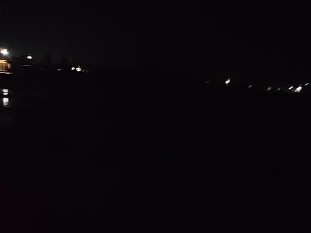 The video captures a dark, nighttime scene, likely from a moving vehicle due to the motion blur. Distant lights are visible, suggesting the presence of buildings or streetlights in the background. The foreground is almost entirely black, with very little detail discernable. The light sources in the background appear as blurred streaks or blobs of light, indicating movement. There are no clearly identifiable people or objects in the frame. The overall impression is one of driving through a dimly lit area at night.