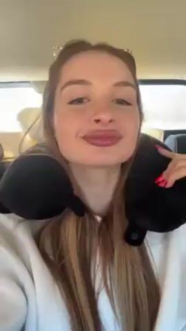 A fair-skinned woman with long blonde hair and pink nails is in a vehicle. She's wearing a white sweatshirt and a black neck pillow. She looks at the camera and smiles, then she starts to talk. Sunglasses are pushed up on top of her head. There are other passengers behind her.