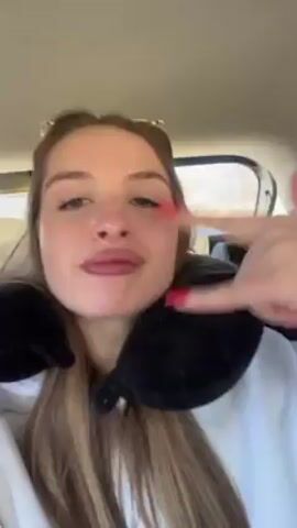 A woman in a car is wearing a neck pillow and sunglasses. She has long brown hair and is wearing a white sweatshirt. She makes a kissy face and winks at the camera. The car window is visible in the background. Music is playing.