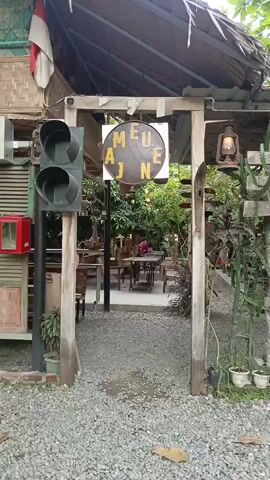 The video pans across the entrance of an outdoor cafe or restaurant. A wooden archway, weathered and rustic, serves as the entryway. To the left of the archway, a vintage traffic light with its red and yellow lights visible stands as decoration. A sign hangs from the archway, a circular metal disc with letters spelling out "MEUE AJN" in a rustic font. Behind the archway, the cafe's interior is visible: a gravel-covered pathway leads to tables and chairs made of wood, set amidst lush greenery and trees. String lights are visible overhead, suggesting an evening ambiance. A person, their back to the camera, is seated at one of the tables, their presence minimal and unobtrusive to the overall scene. The environment appears relaxed and natural, with a blend of industrial and organic elements in its decor.