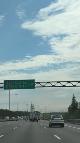 The video shows a view from a car driving on a multi-lane highway. The sky is blue with white clouds. A green highway sign is visible overhead, indicating that "KOLA" is 5.8 kilometers away. Several other vehicles, including cars and trucks, are on the highway in front of and beside the camera's vehicle. Trees and power lines are visible along the sides of the road.