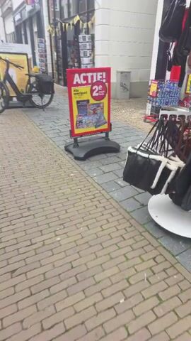 The video shows a pedestrian street in a European city. The street is paved with bricks, and there are shops on both sides. In the foreground, a bicycle is parked on the sidewalk. Further down the street, there is an ice cream shop with red benches outside. There are a few people walking on the street, and some shops have promotional signs outside. The weather appears to be overcast.