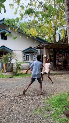 The video shows three men playing a game in a gravel yard next to a white building with blue-trimmed windows and a small wooden pergola with seated people inside. The man in the center, wearing a light blue t-shirt and dark shorts, kicks a brown, round object high into the air. The camera follows the object as it’s passed to a man in a green t-shirt and dark shorts who jumps and hits it even higher. It sails over the white building. The men laugh as it flies over the roof. The scene is set outdoors during daylight hours and trees with yellow flowers are visible.