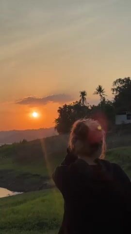 A vertical video shows a young person with blonde hair tied up in a bun, wearing a black hooded sweatshirt, standing in a grassy area overlooking a body of water and rolling hills at sunset. The person is facing away from the camera, with their right arm raised and hand touching their head. The sun is visible in the sky, casting an orange glow and lens flares across the scene. Trees and some distant structures are visible on the hills. The water is calm, reflecting the sky. The grass is green and appears to be slightly sloped. The overall ambiance is peaceful and serene.