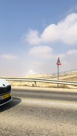 A roadside view shows a desolate landscape with a bright blue sky and scattered clouds. In the foreground, a paved road runs horizontally, with a guardrail separating it from a dirt and scrubland area. A triangular warning sign with a curved arrow indicating a sharp turn is visible to the right of the road. In the background, a tall, slender tower stands prominently, emitting a bright beam of light or steam into the hazy sky, suggesting a solar power plant or similar industrial facility. The overall impression is one of an arid, possibly desert environment with a man-made structure dominating the distant horizon.