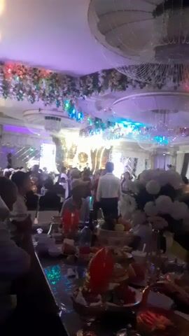 The video shows a crowded event hall during what appears to be a celebration or reception. The hall is brightly lit with purple and white lighting, and decorated with floral arrangements and hanging lights. People are seated at tables laden with food and drinks, and others are standing and socializing. In the background, there is a stage area with large screens and decorative elements, suggesting a performance or presentation is taking place. Some individuals are visible dancing or moving around the hall. The overall atmosphere seems festive and lively.