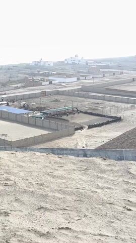 The video presents a wide-angle, slightly hazy view of a sandy landscape with signs of construction. In the foreground, a large expanse of sand is visible with visible tracks or patterns on the surface. Behind the sand dune, there is a construction site surrounded by concrete block walls. Within the site, there appears to be a structure covered with a blue tarp, and another structure supported by posts. There are dark, possibly rubber objects lined up in a semi-circular fashion at the edge of the site. Further in the background, a settlement or residential area with several buildings is discernible. The buildings seem to be predominantly white or light-colored and vary in size and design. The sky is overcast, contributing to the hazy atmosphere. The overall impression is that of an arid or coastal environment with ongoing construction or development activity. No individuals are readily visible in the video.