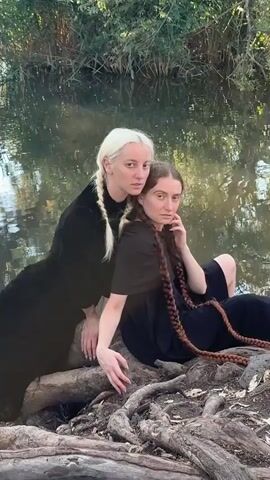 Two women, one with blonde hair in a braid and wearing a black dress, and another with brown hair in a long braid and wearing a black dress, pose near a body of water. The blonde woman leans over the seated brown-haired woman, placing a hand on her arm. The brown-haired woman rests her head slightly back and touches her neck with her other hand. They are positioned on logs and tree roots near the water's edge, with dense foliage in the background.