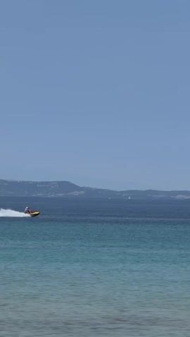 A wide shot shows a jet ski speeding across the water, leaving a white spray behind it. In the foreground, the shallow water laps against a sandy beach, and several people are wading or swimming. The background features a distant landmass, possibly a coastline, under a clear blue sky. The scene is peaceful and evokes a sense of summer leisure.