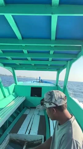 A person wearing a camouflage cap and a gray t-shirt is seated inside a boat, facing forward and to the right. The boat has a turquoise exterior and a blue ceiling supported by turquoise beams. The interior of the boat features turquoise benches and wooden flooring. The camera is positioned to capture the scene from behind the person, showing the boat moving through the water. The ocean is visible to the right, with waves and foam trailing the boat. In the distance, a hazy landmass with buildings can be seen on the horizon under a clear blue sky. Another small boat is visible in the distance. The video appears to be shot during the daytime.