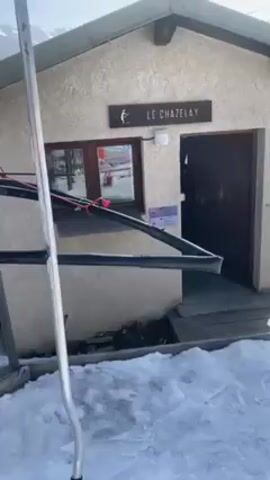 The video shows a ski lift moving away from a small building at a ski resort. The building has a sign that says "LE CHAZELAY".  The ground is covered in snow and a few people can be seen skiing in the distance. The ski lift's poles are visible in the foreground. The sky is clear and blue.