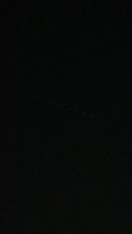 A line of small white lights appears to move across a completely dark sky. There are background sounds of people speaking.