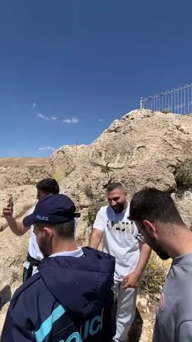 The video shows a group of men, one wearing a police jacket, standing on a rocky hilltop overlooking a town in what appears to be a desert landscape. The camera pans to reveal a statue on the hill, and a view of the town and surrounding landscape. The sky is blue with scattered clouds. The men are talking and interacting with each other.