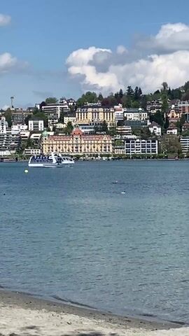 The video shows a sunny day on Lake Lucerne, Switzerland. A large white tour boat is moving across the water. In the background, a hillside is covered in various buildings, including the Montana and Palace hotels. The sky is blue with scattered white clouds. The shoreline in the foreground has a sandy beach. The water is calm with a few small ripples.