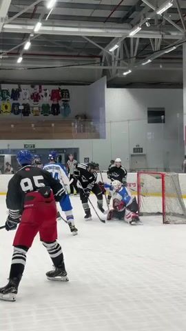 A hockey game is taking place on an indoor ice rink. Several players are on the ice, some wearing blue and white uniforms, and others in black and red. A player in a black jersey with the number 95 on the back and red pants is in the foreground, facing away from the camera. In the middle of the ice, a scrum is occurring around the net. One player is on the ice, and others are trying to gain possession of the puck. A goaltender in full gear is positioned in front of the net. The arena has a high ceiling with exposed metal beams and lighting fixtures. Posters of hockey jerseys are displayed on a wall in the background.