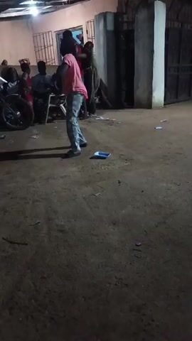 A person in a pink shirt and patterned pajamas is dancing in a dirt yard at night. They are moving their arms above their head. There are people watching, some sitting in chairs and others standing. A motorcycle is parked nearby. There is trash scattered on the ground and buildings in the background.
