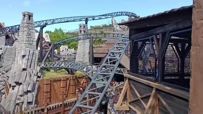 The video shows a roller coaster with a gray track that goes over a bridge and through rocky structures. A roller coaster train with people on it is visible on the track. The scene appears to be an amusement park with a clear blue sky and some green foliage in the background.